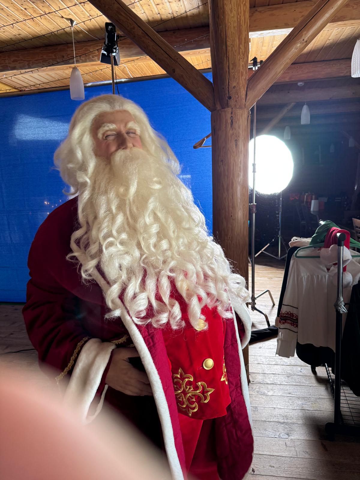 Święty Mikołaj z profilu na planie - niebieskie tło, lampa filmowa i stojak z kostiumami